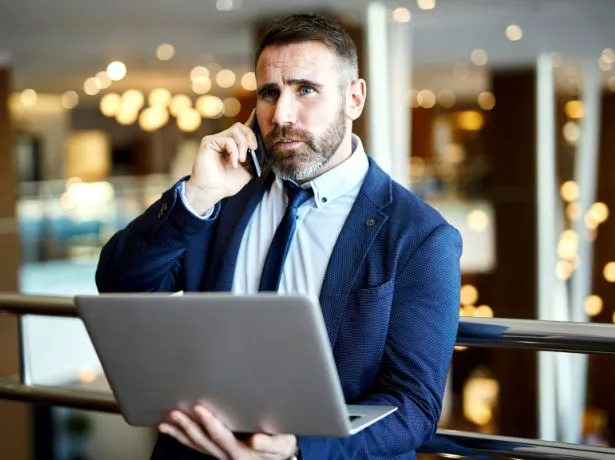 Imprenditore con la barba e giacca blu al telefono con un computer portatile in mano